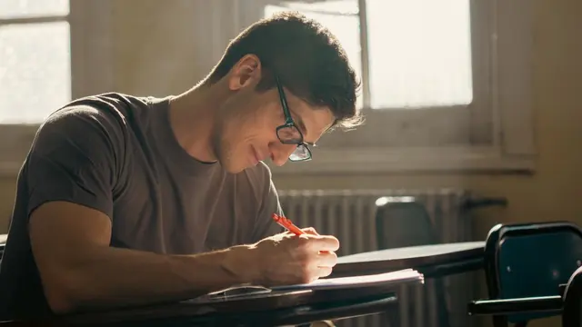 Student in glasses writing answers at a desk by a window