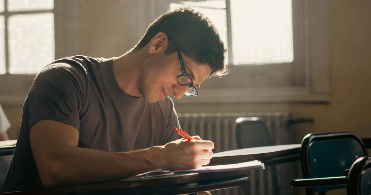 Student in glasses writing answers at a desk by a window