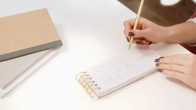Hands writing notes in a spiral notebook on a white desk