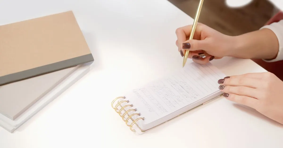 Hands writing notes in a spiral notebook on a white desk
